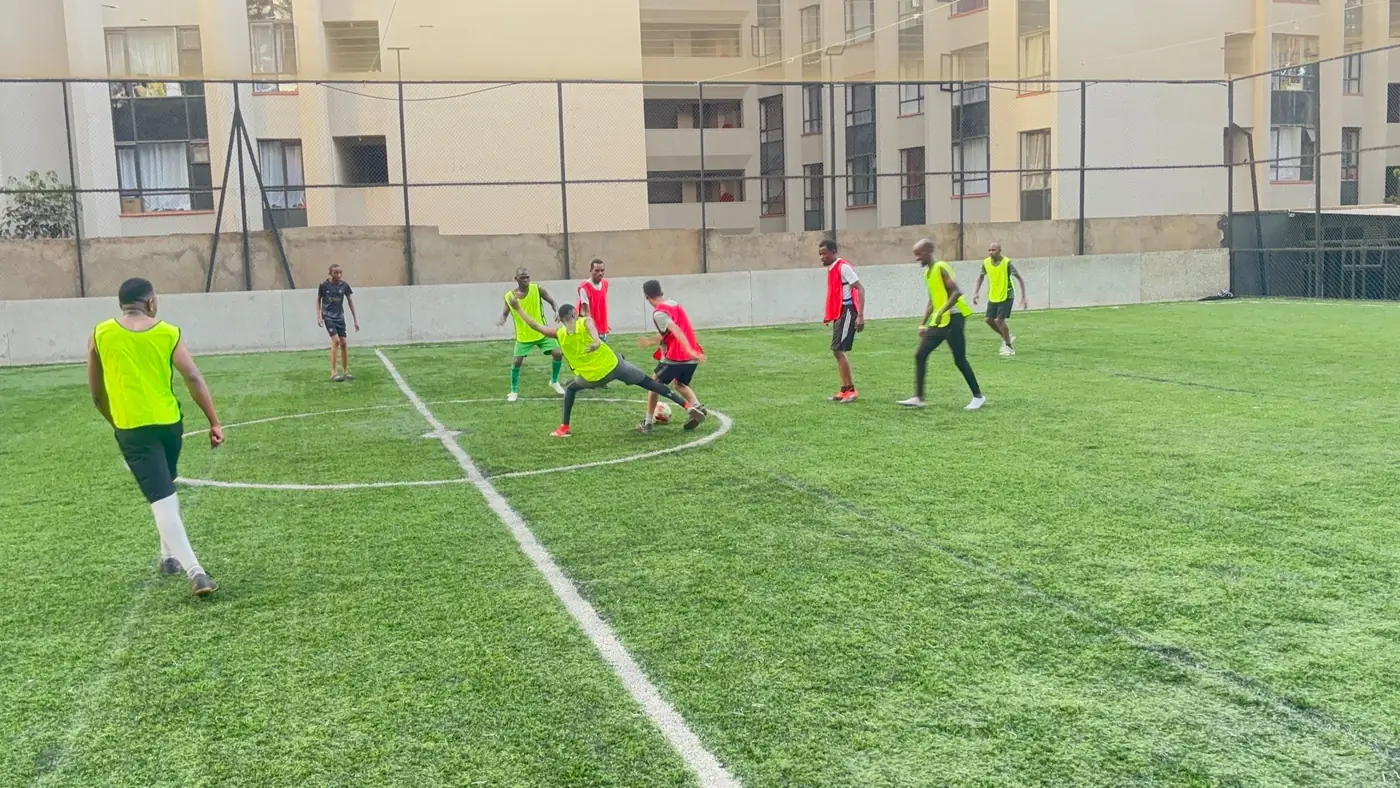 Players in yellow and red bibs fight for the ball in the centre circle of an artificial pitch, with flats rising behind the fence.